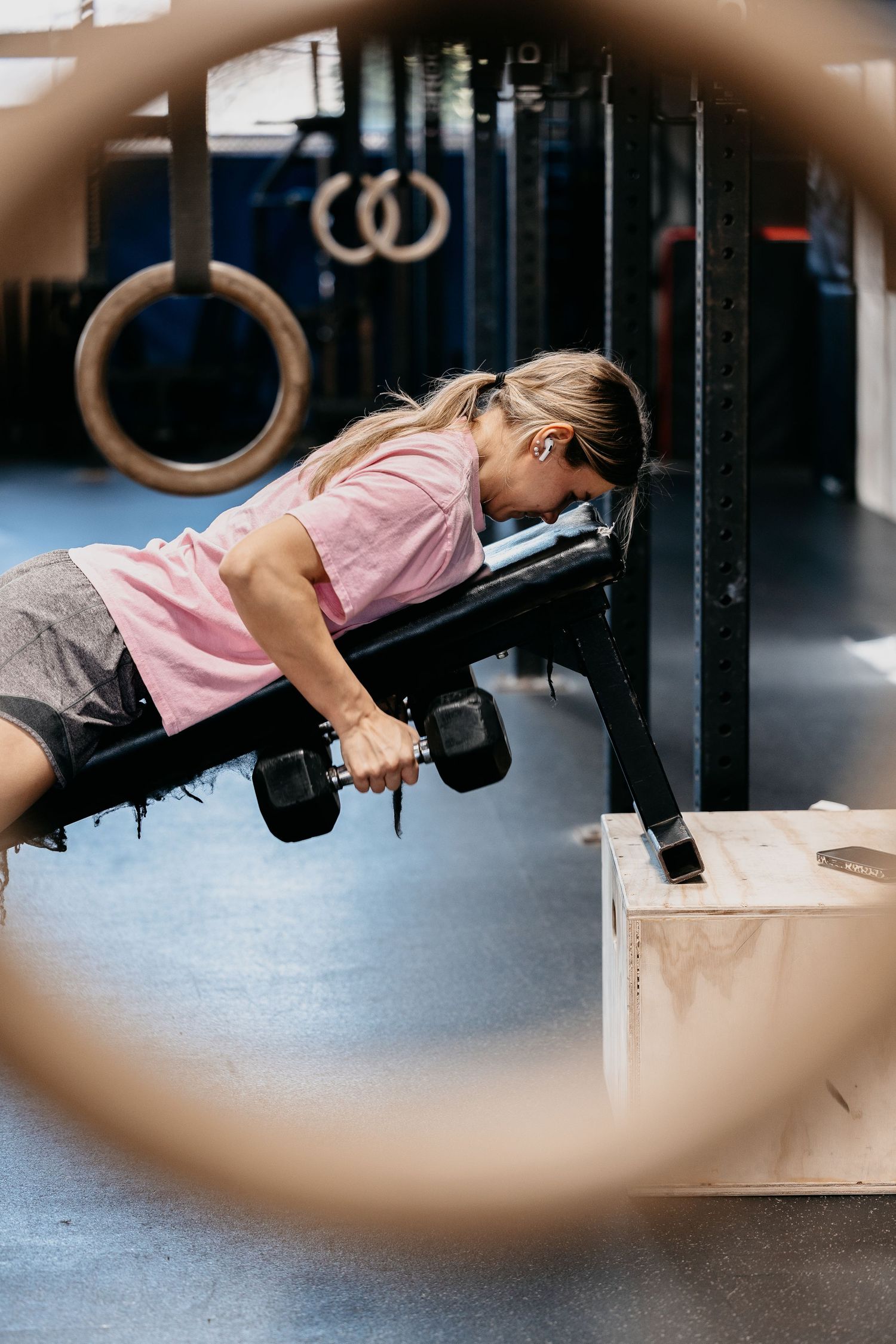 Woman in pink shirt doing dumbbell rows on a bench in a CrossFit gym with gymnastic rings in background.