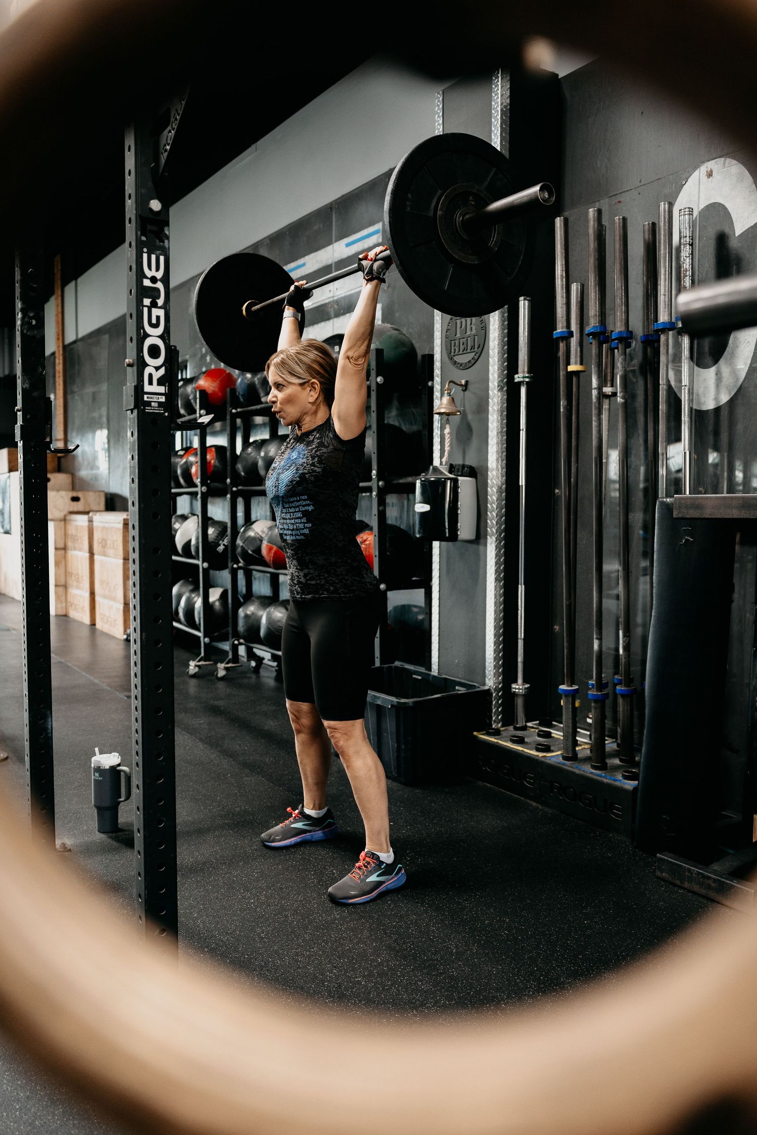 Woman performing overhead barbell press in a Rogue squat rack at a CrossFit gym wearing black athletic wear.