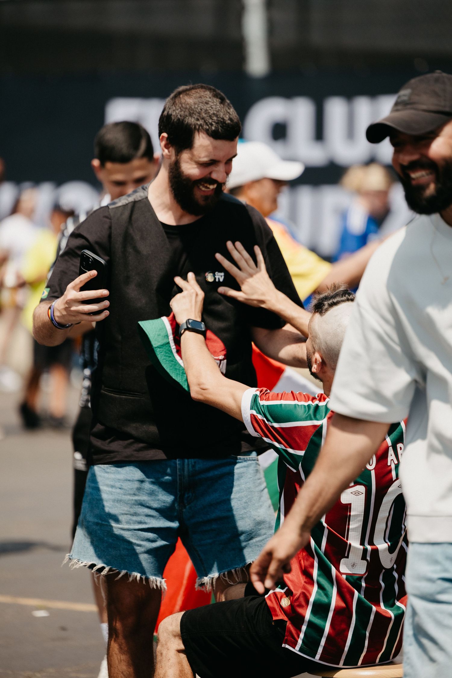 Bearded man in black tee laughing with friends outdoors at a sunny street gathering.