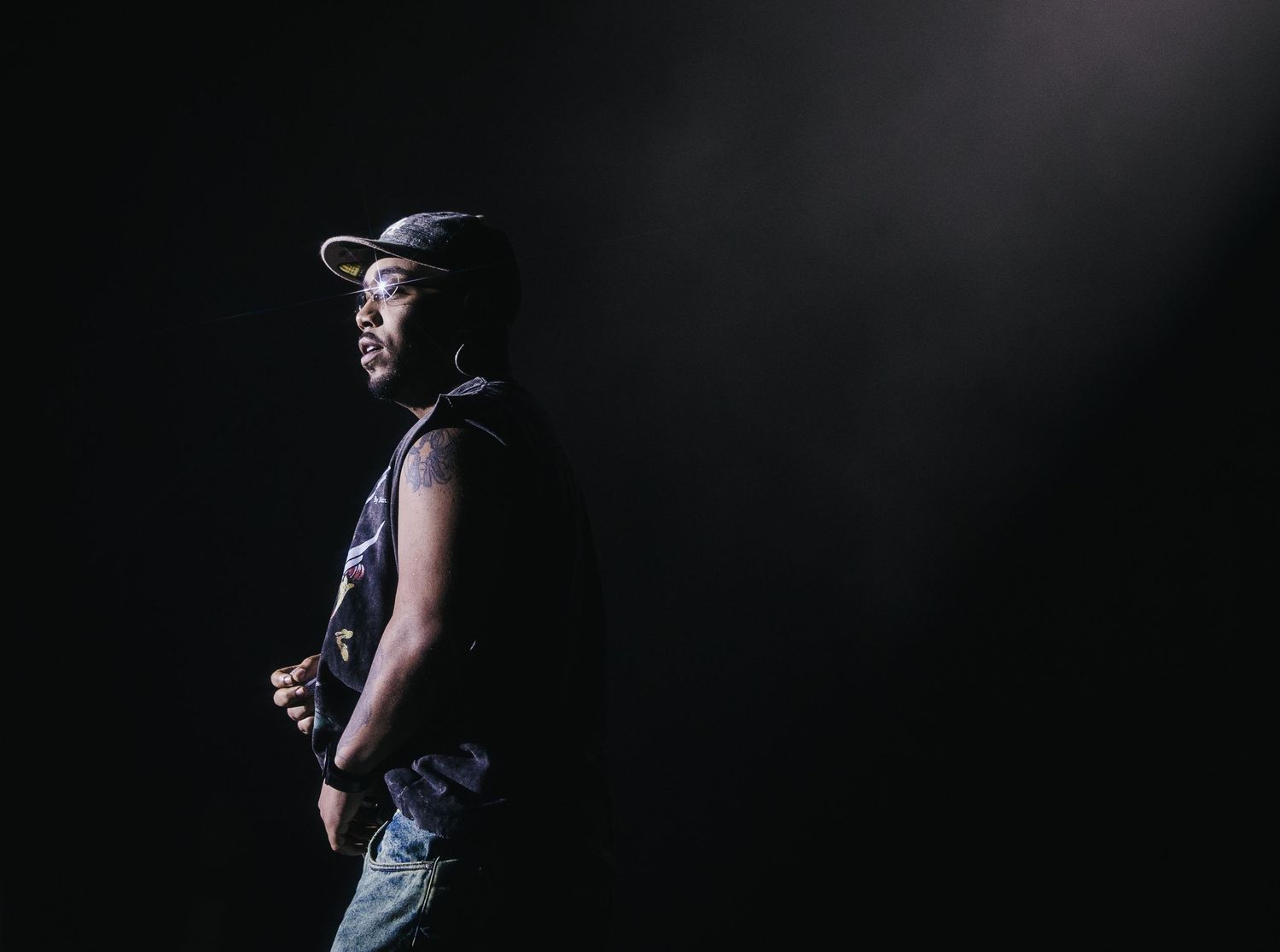 Musician stands in dramatic spotlight against a dark background wearing a sleeveless shirt and cap.