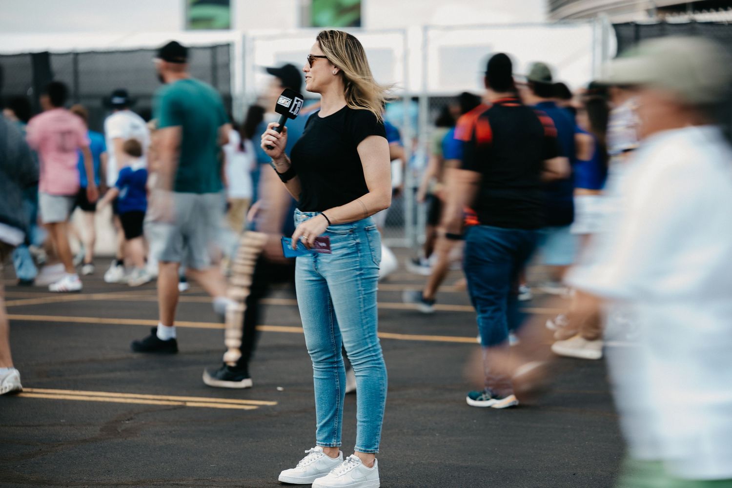 Woman in black top and jeans standing still amid blurred crowd at outdoor event.