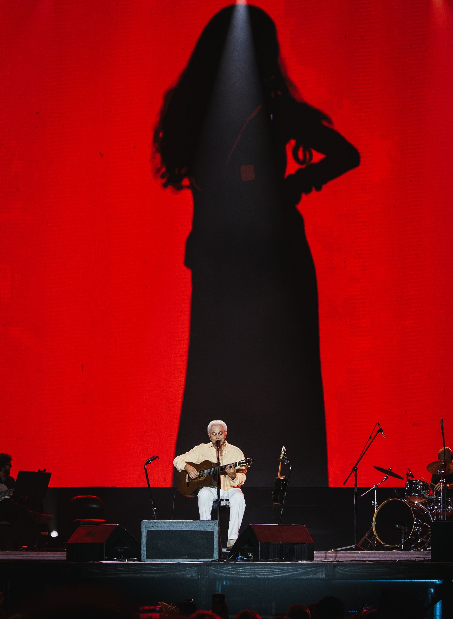 Guitarist performs solo on stage beneath a dramatic red backdrop with a large female silhouette shadow projection.