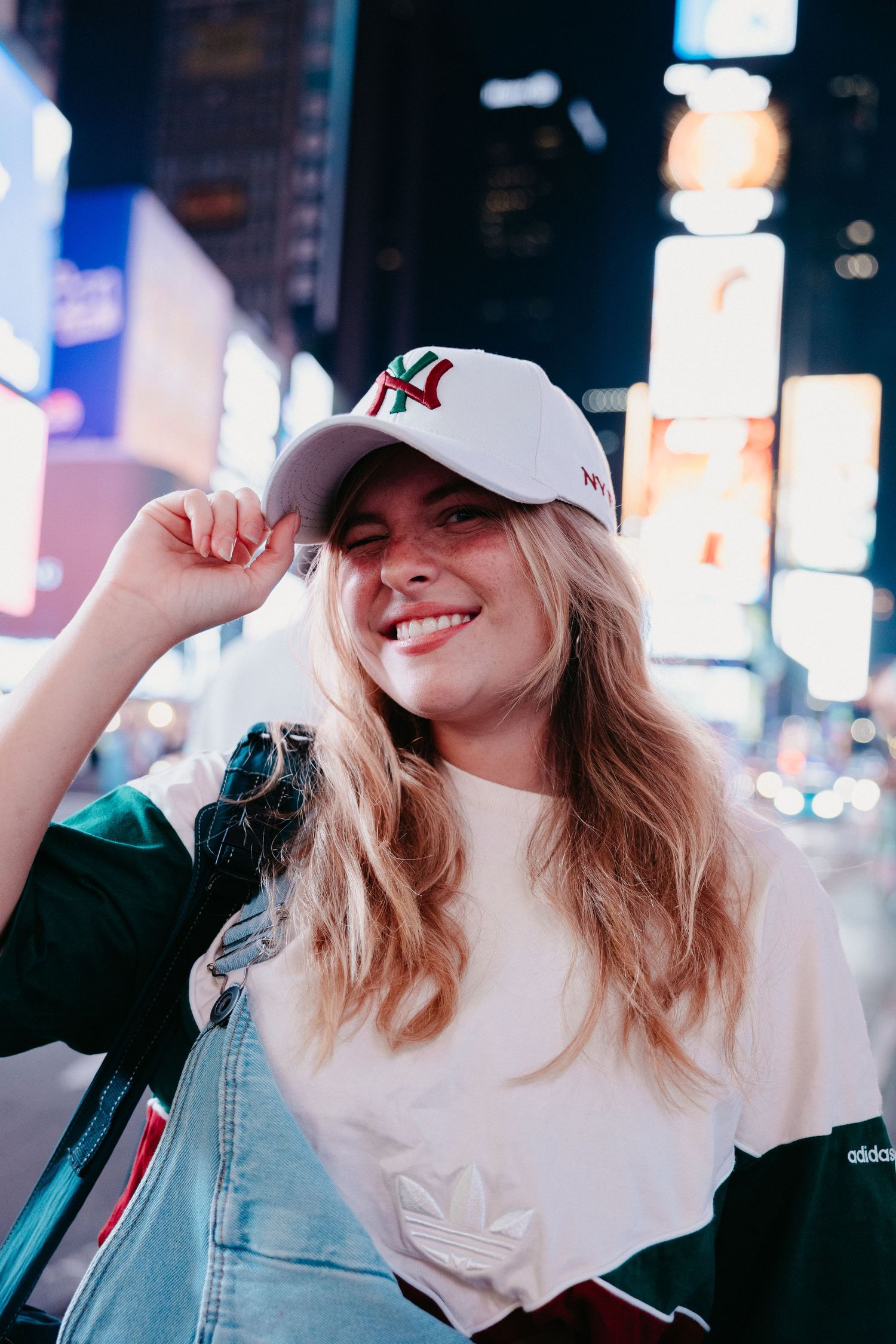 Smiling blonde woman adjusting NY Yankees cap on Times Square street at night with city lights.