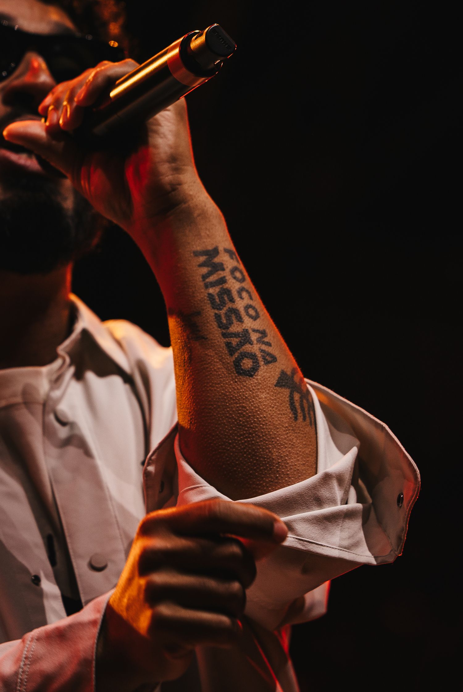 A performer holds a microphone revealing a forearm tattoo reading 'Foco na Missao' during a live show.