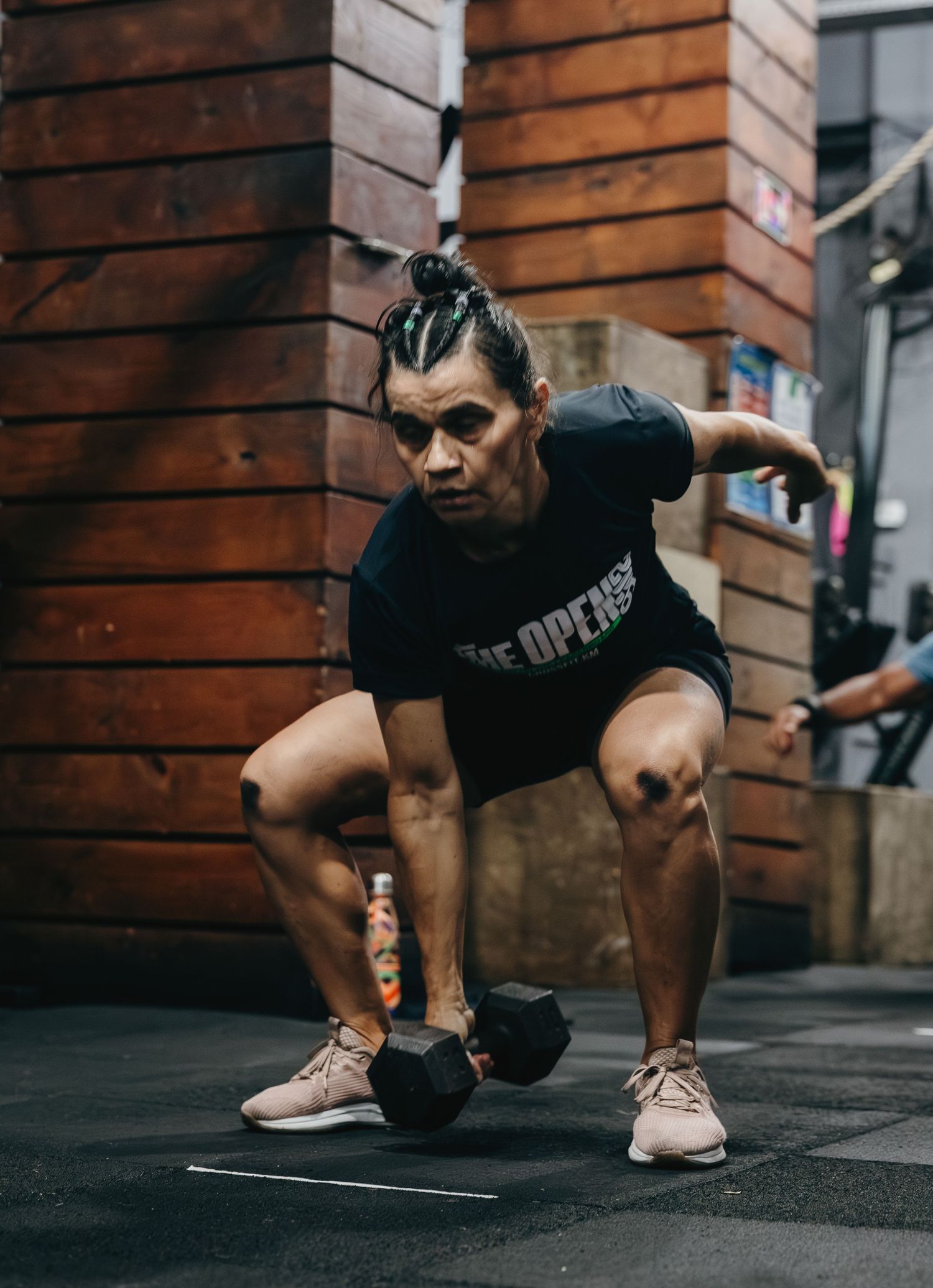 Athlete in black t-shirt performs dumbbell squat exercise on gym floor with focused expression.