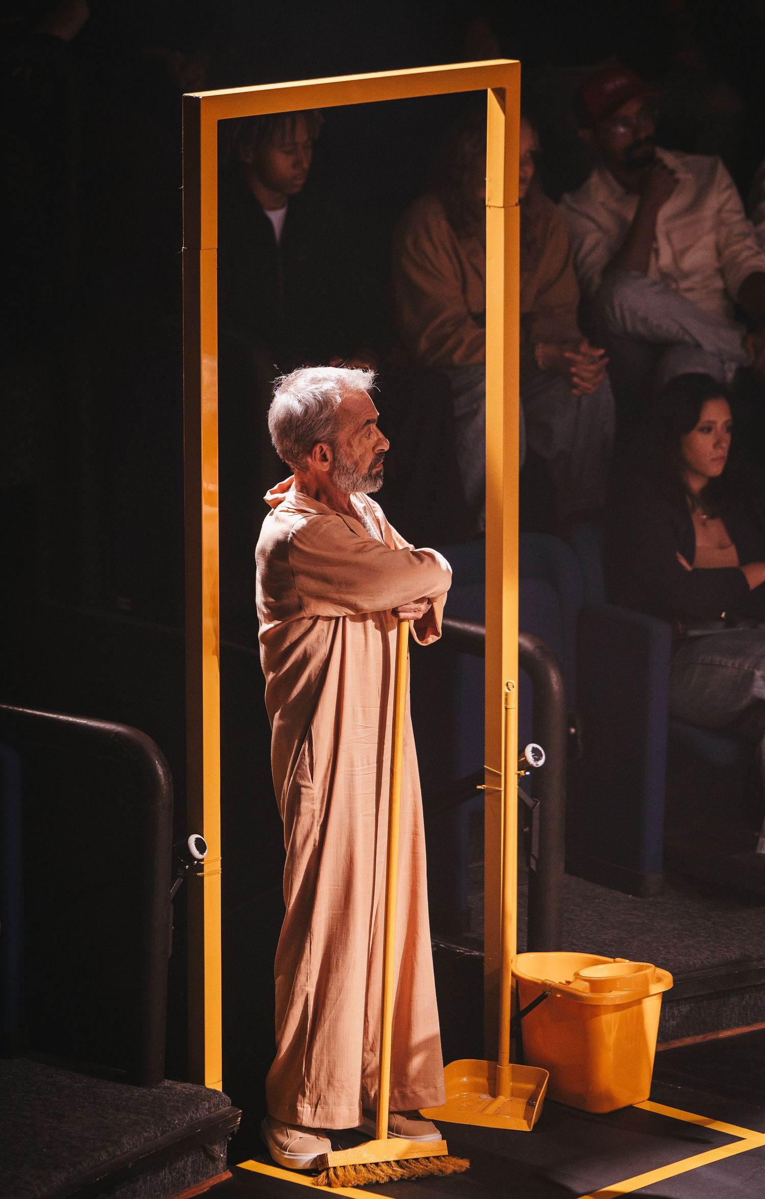 Elderly man in a beige robe stands with crossed arms inside a yellow door frame on a theater stage.