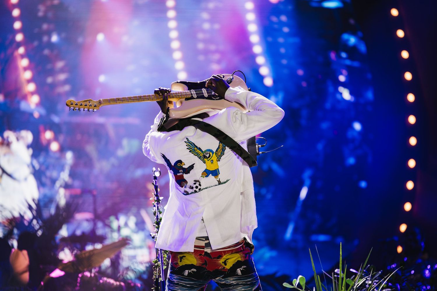 Guitarist performing on stage in a white graphic jacket, holding guitar overhead under colorful concert lights.