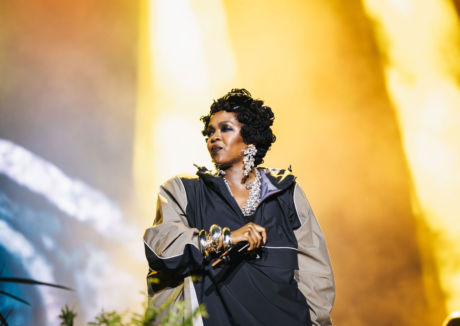 Female performer on stage in black outfit with statement jewelry, singing under dramatic golden stage lighting.