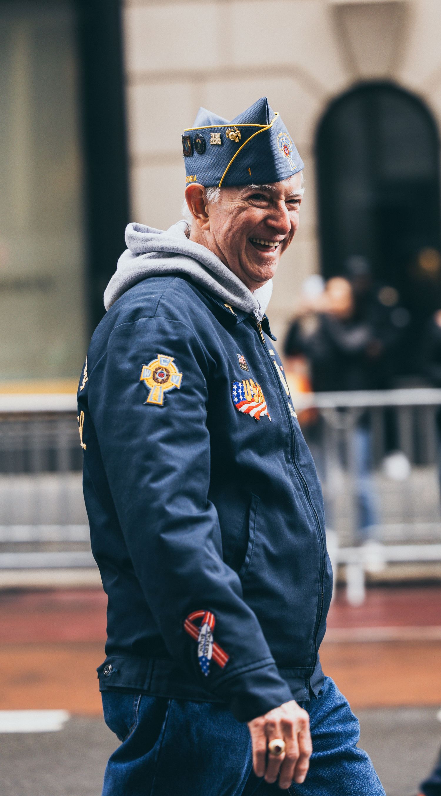 Smiling veteran in navy jacket with patches and garrison cap walking in an outdoor parade.