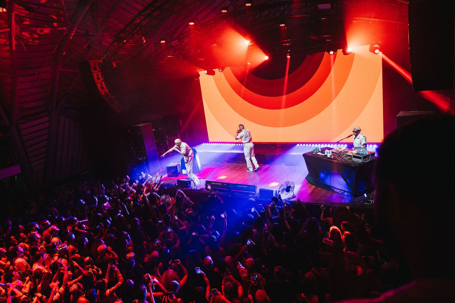 Energetic concert scene with performers on stage before a large crowd under colorful lights.