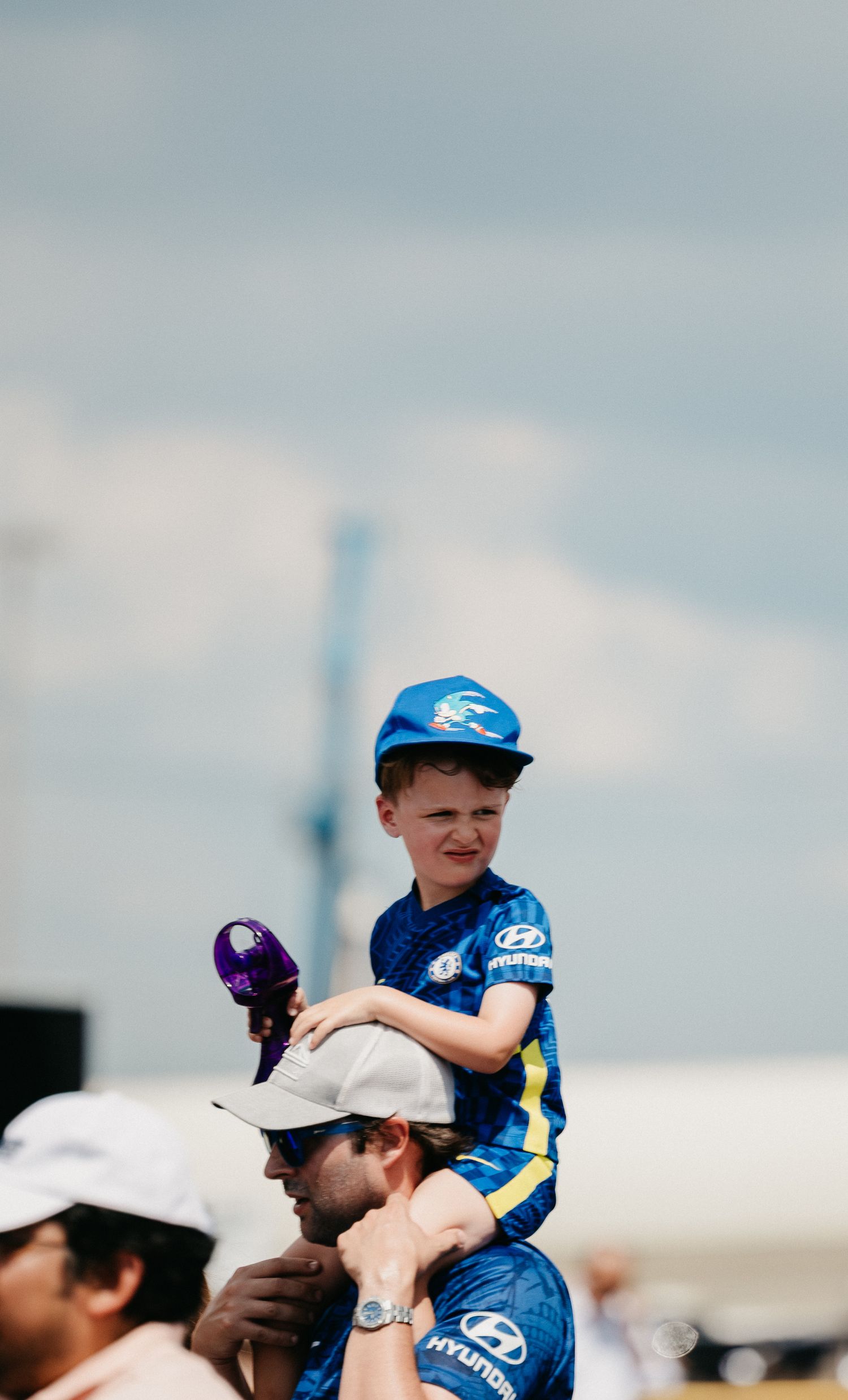 Young boy in blue Hyundai jersey and helmet sitting on mans shoulders at an outdoor event.