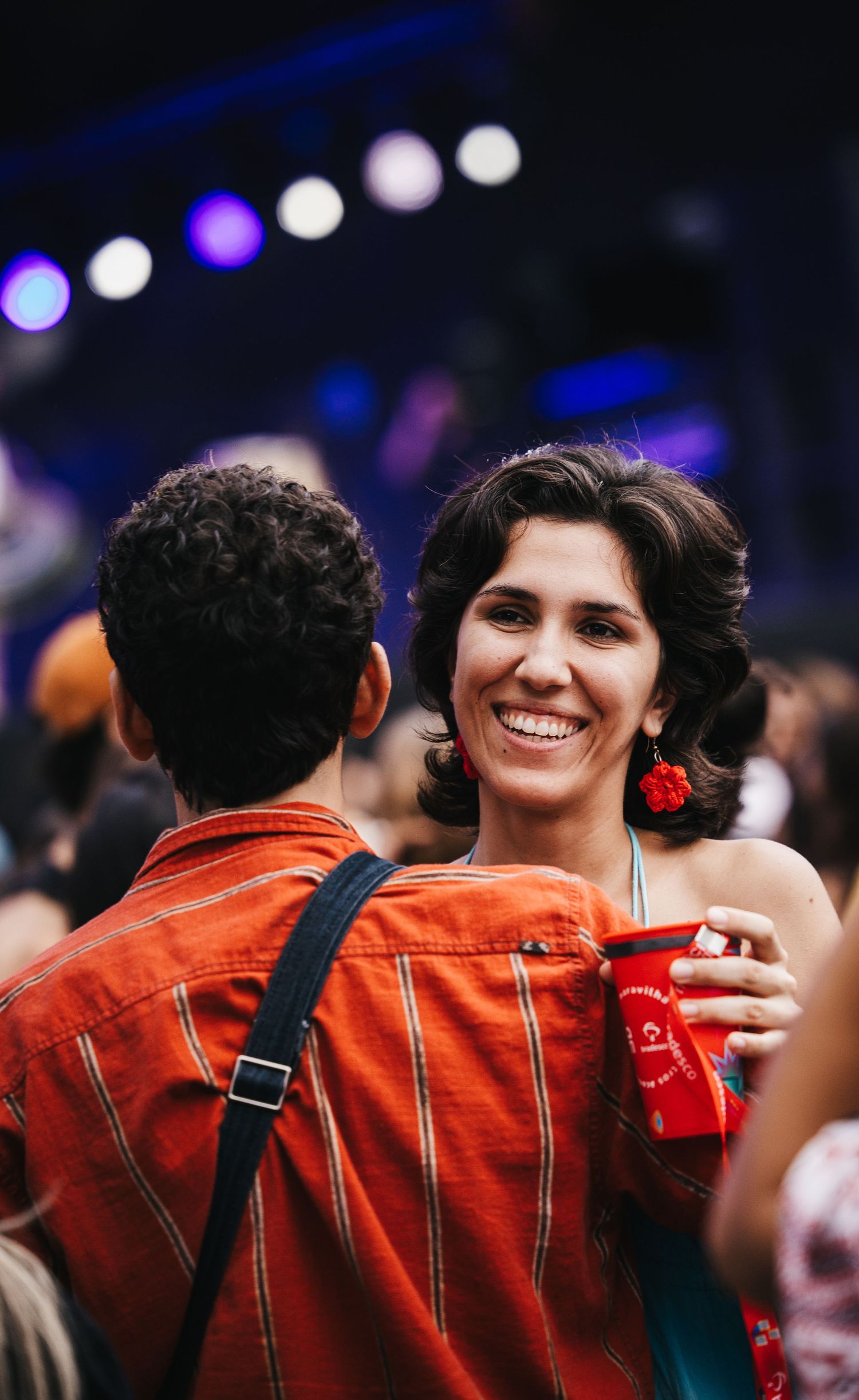 Smiling woman with red earrings holding a red cup at a vibrant concert event with colorful bokeh lights.