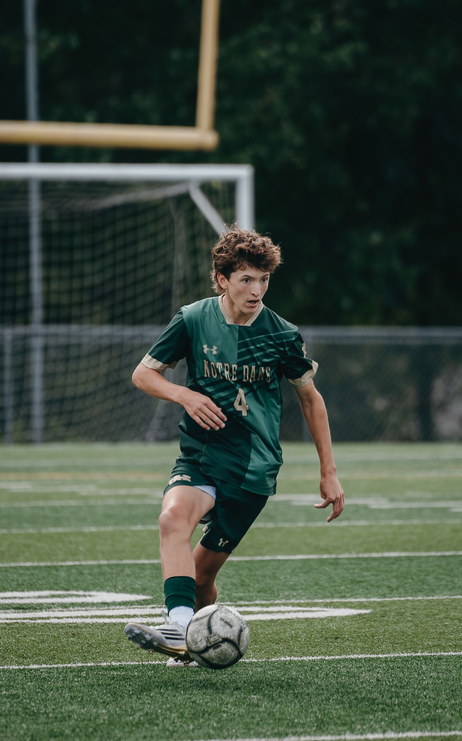 A young soccer player in green uniform dribbles a ball on a turf field during a game.