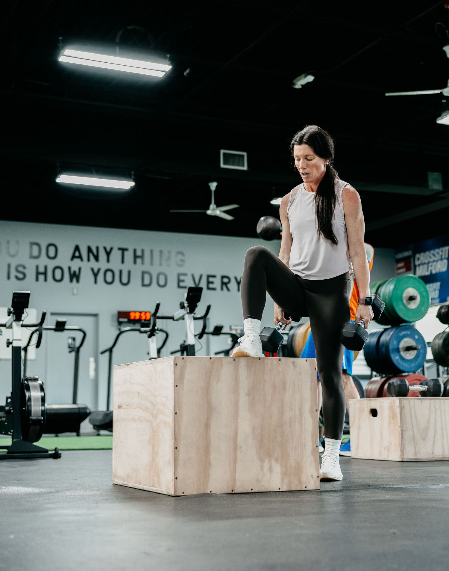 Woman performing box step-ups with dumbbells in a CrossFit gym, wearing black leggings and white sneakers.