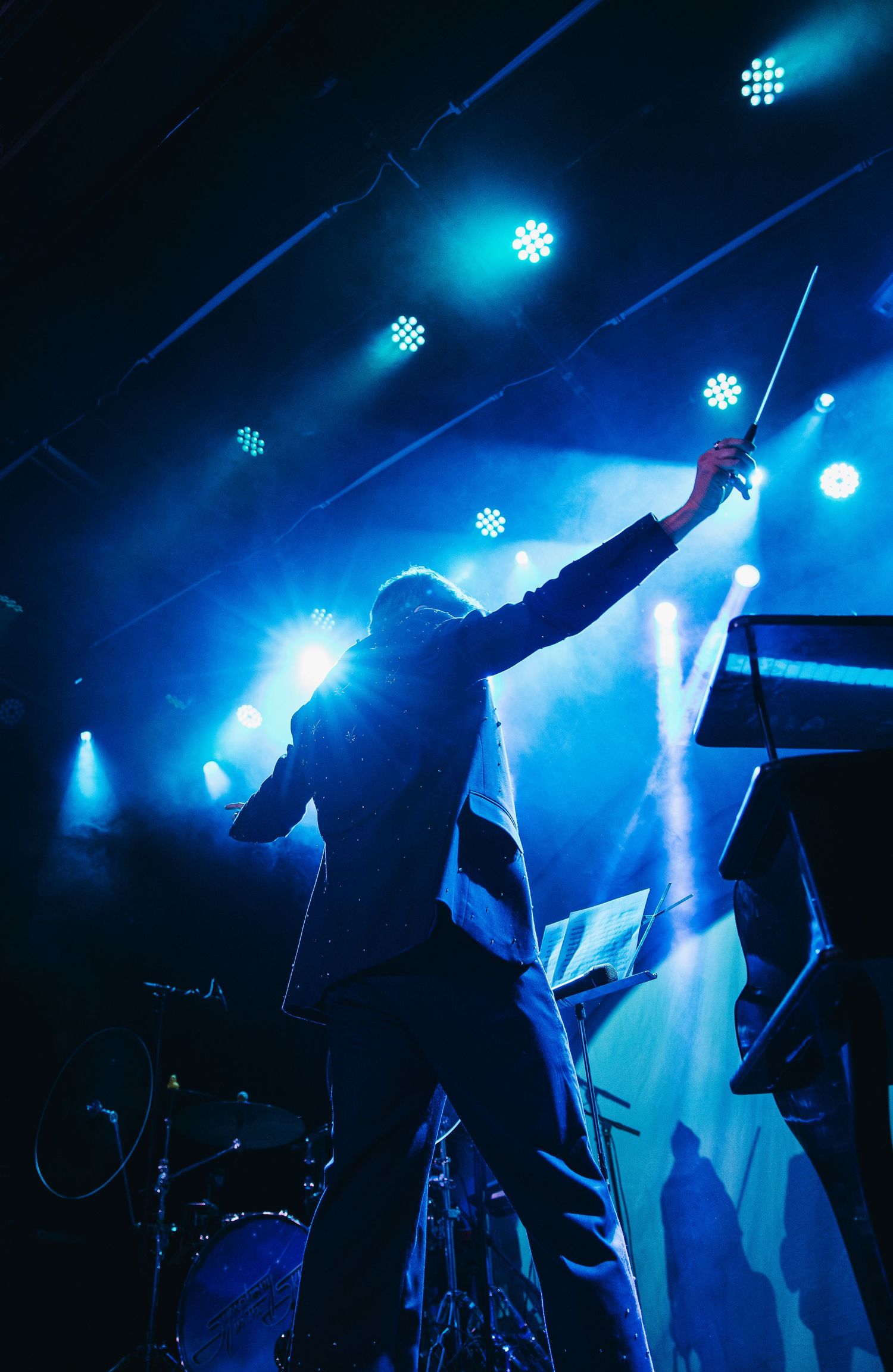 Performer raises a baton on stage surrounded by blue concert lights and stage equipment.