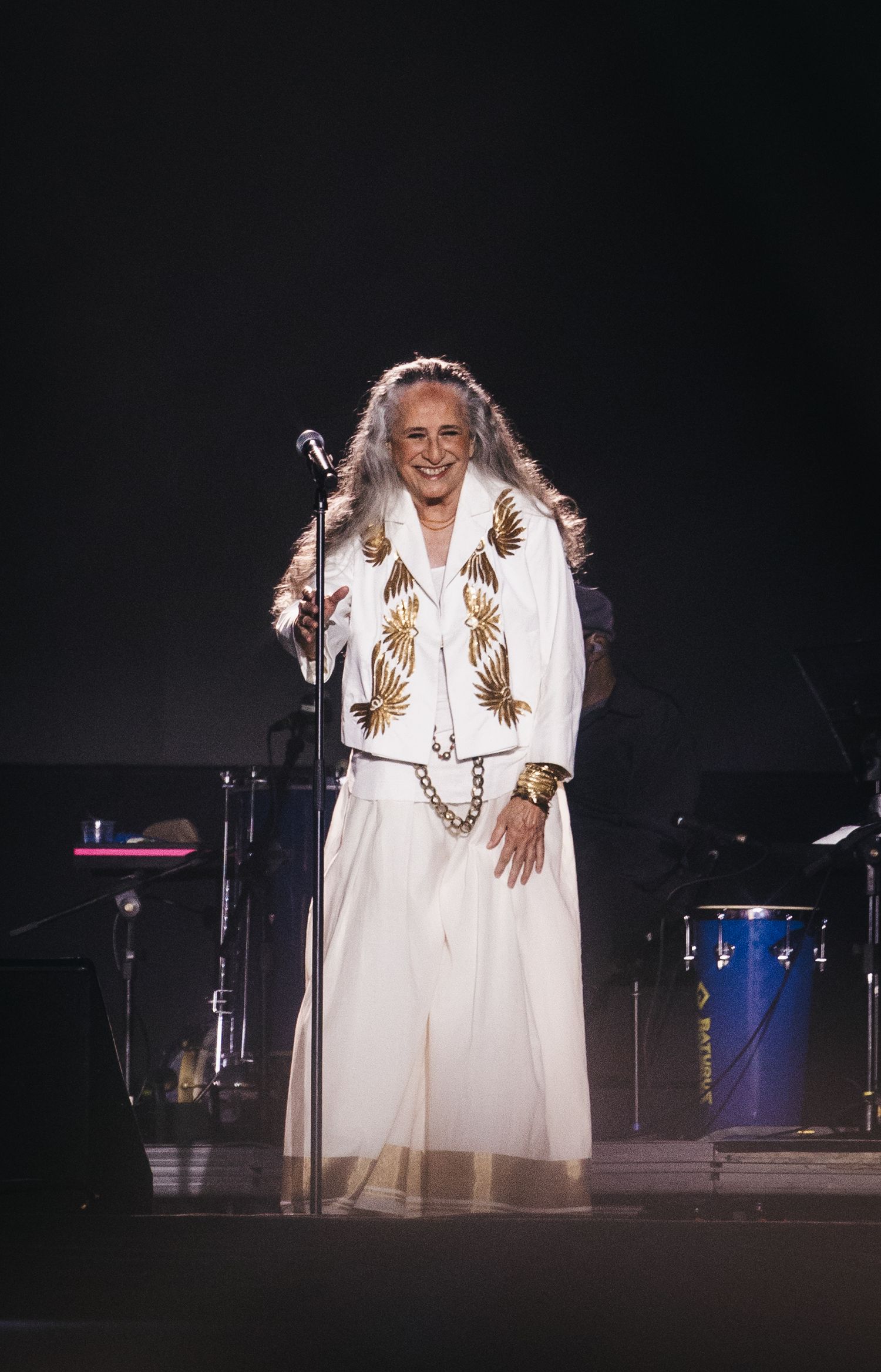 Elderly woman in white and gold embroidered outfit performing on stage at a live concert with microphone stand.