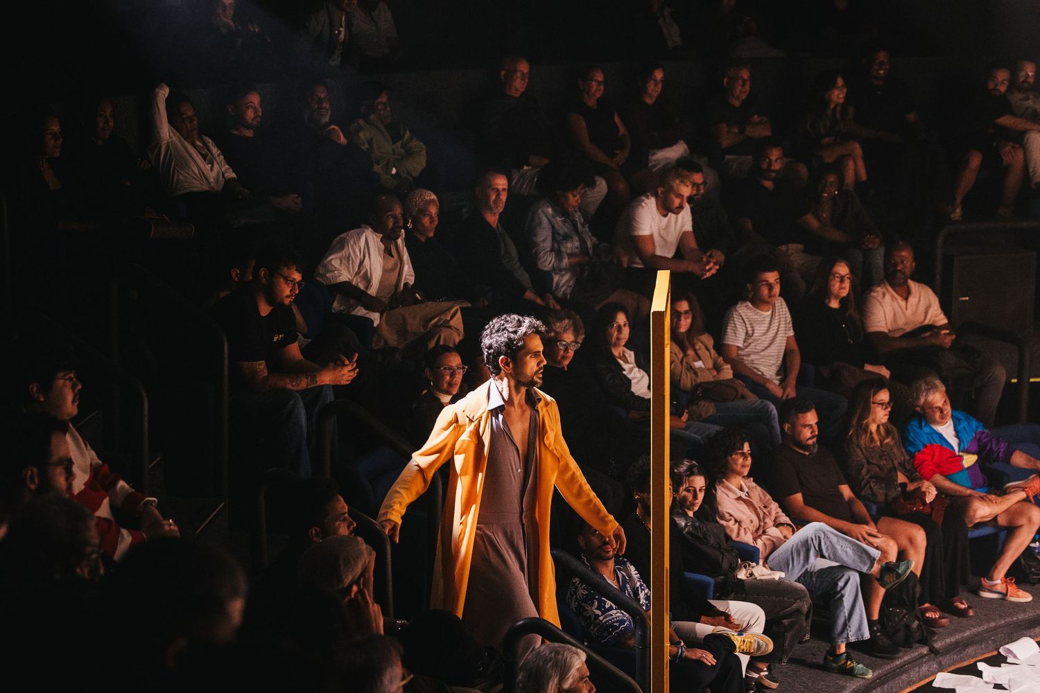 Model walks the runway in a yellow coat at a fashion show, surrounded by a diverse seated audience in a dimly lit venue.