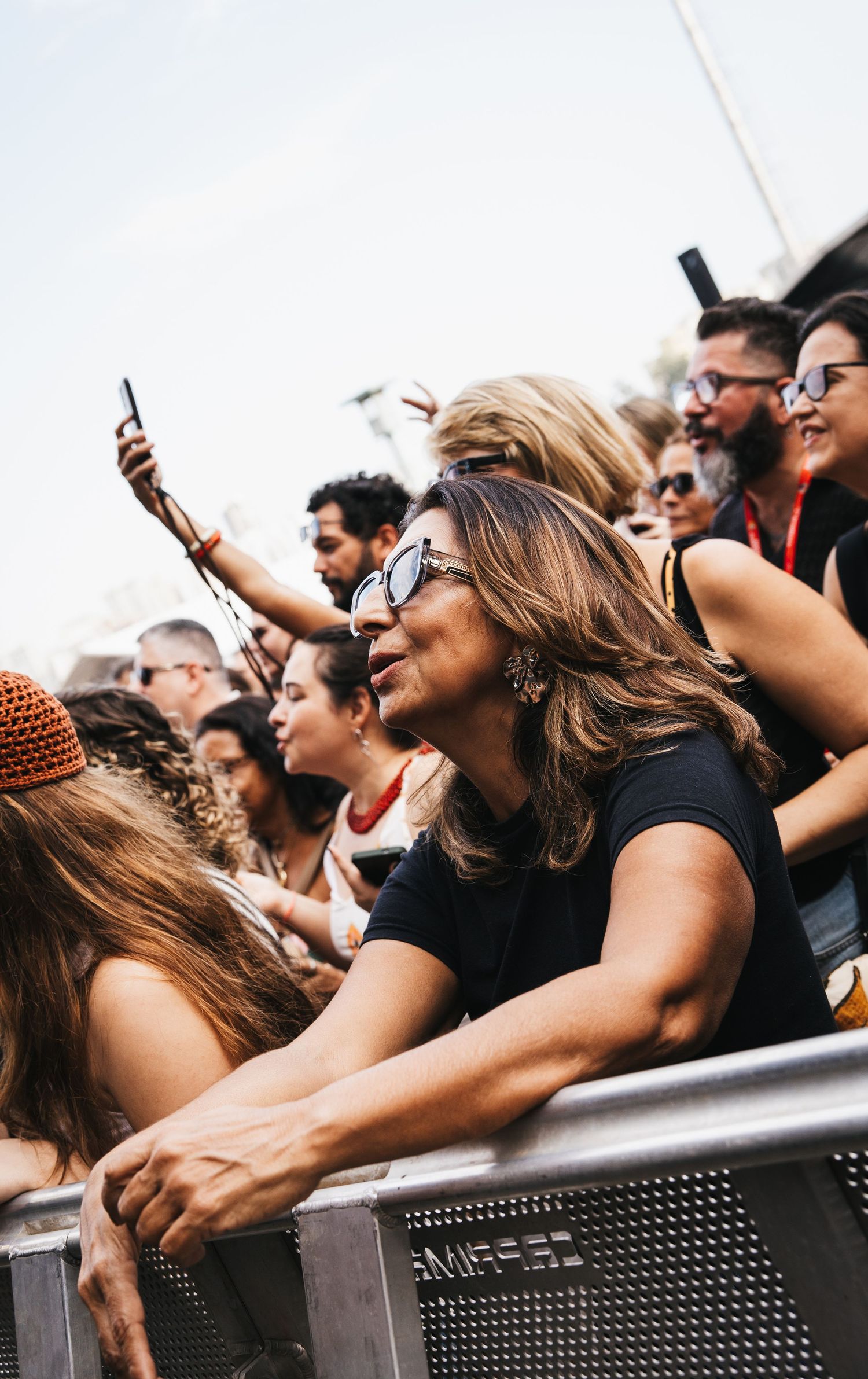 Crowd of diverse concert-goers leaning on barrier, woman in sunglasses smiling at outdoor music festival.