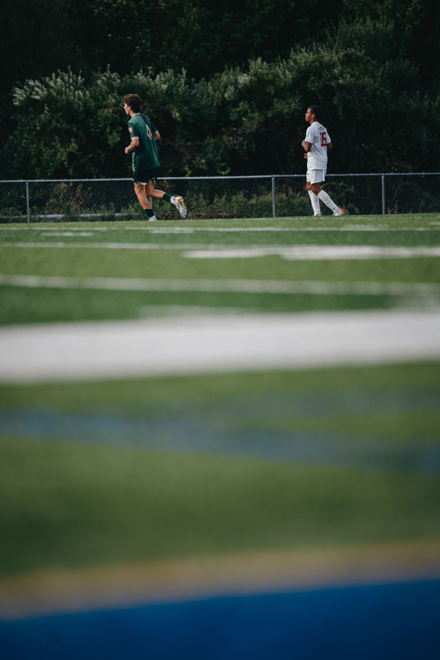 Two soccer players on a green field, one in green kit and one in white, during an outdoor match near a fence.