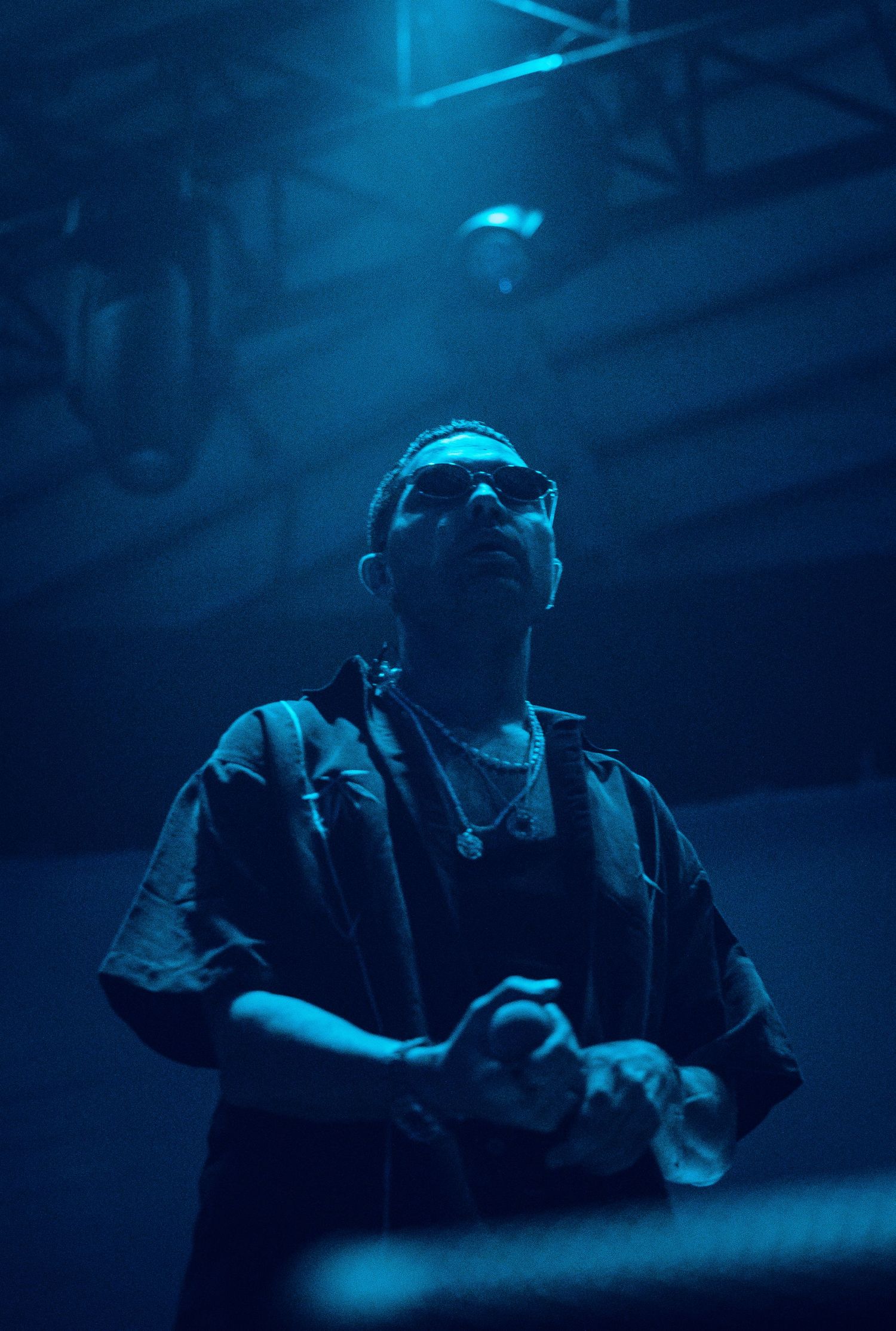 Male artist performing on stage under blue atmospheric lighting wearing sunglasses and dark outfit.