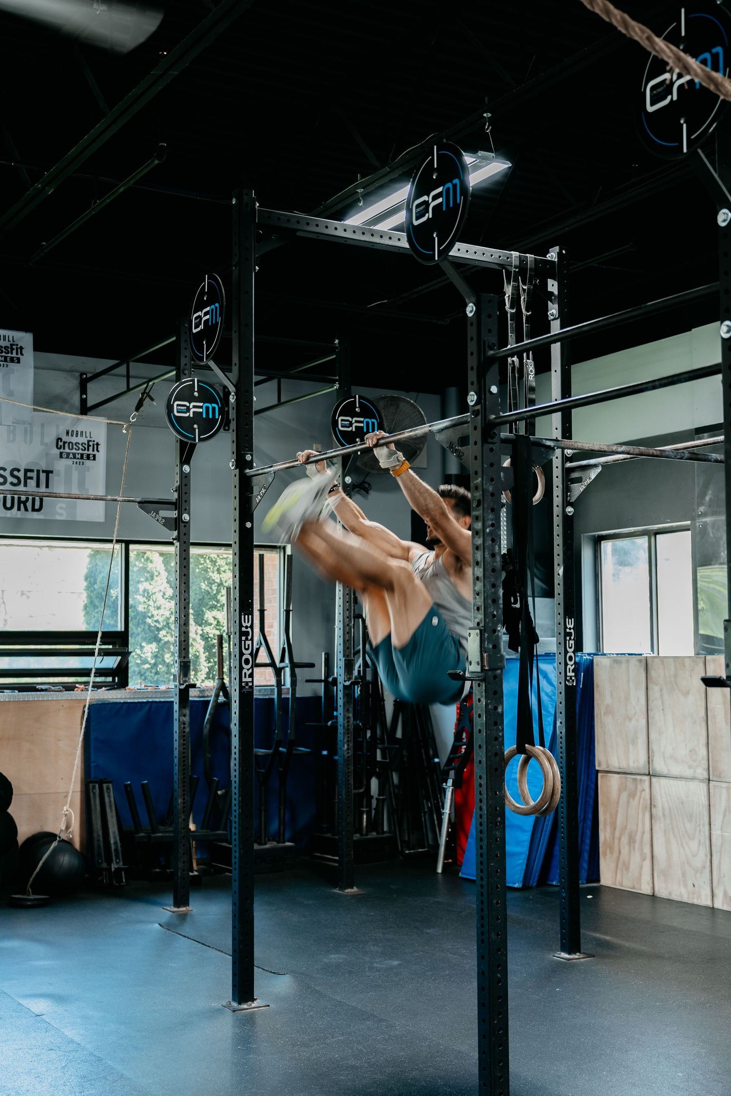 Athlete performing dynamic pull-ups on a rig in a CrossFit gym with gymnastic rings and training equipment.