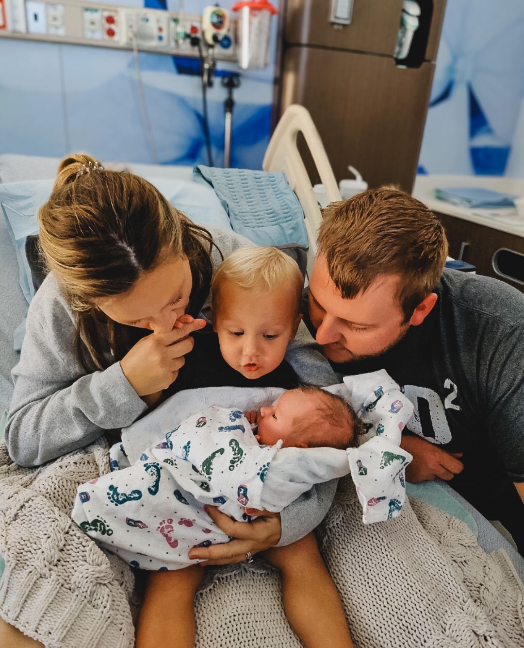 Siblings sitting together on a bed while holding their newborn baby in a cozy lifestyle newborn moment