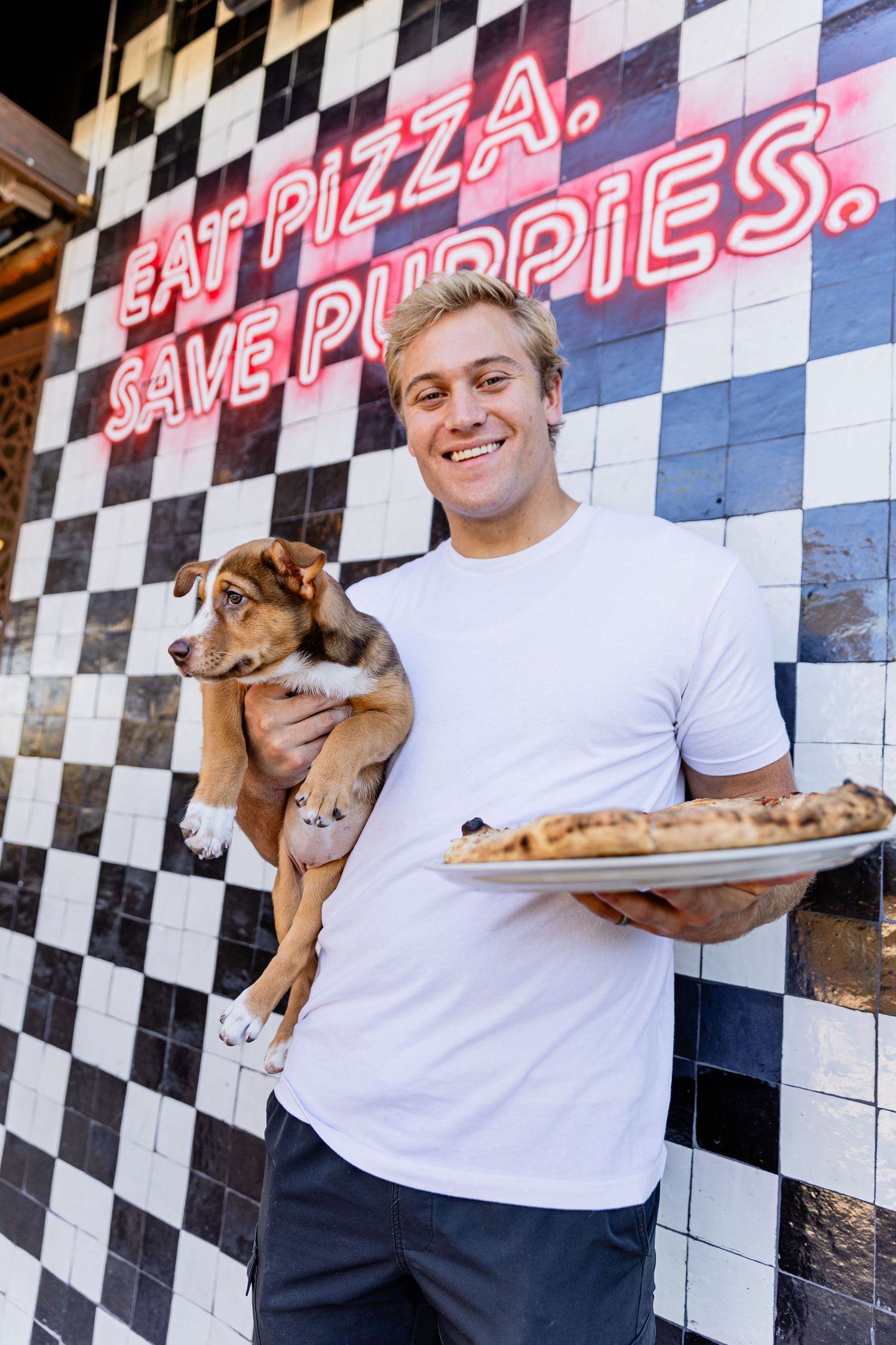 Smiling man holding a puppy and pizza in front of a checkered wall with neon sign reading Eat Pizza Save Puppies.