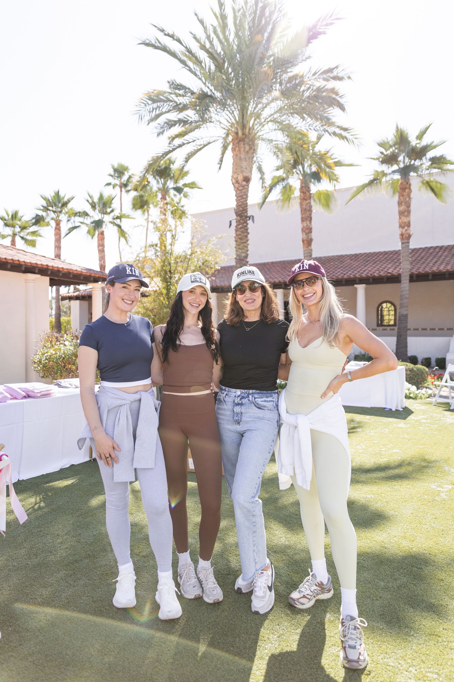 Four women in athletic wear and baseball caps pose together at an outdoor event with palm trees.