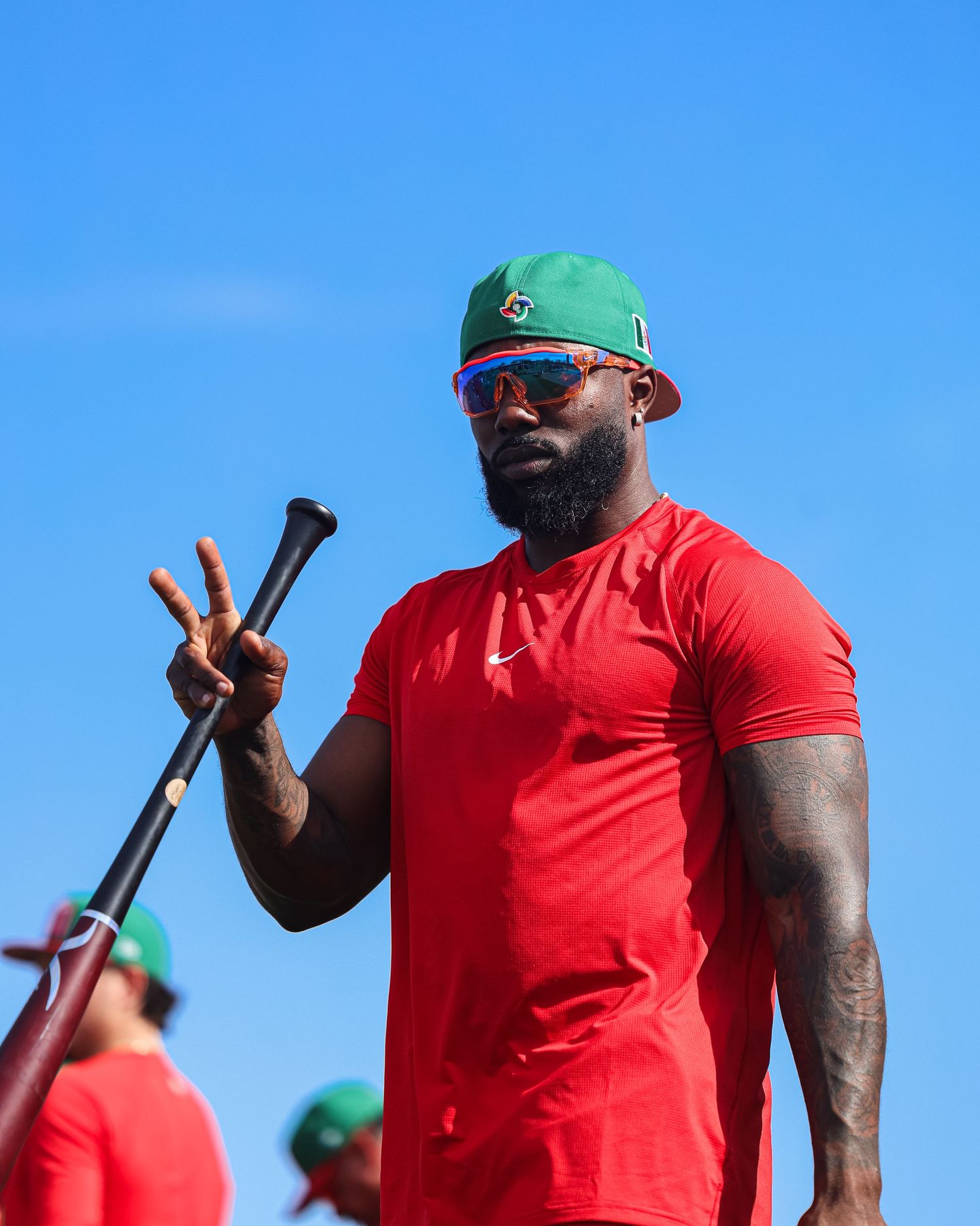 Baseball player in red Mexico uniform holds a bat and flashes a peace sign outdoors.