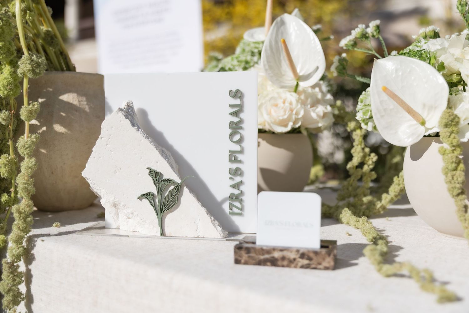 Izas Florals branded sign displayed on a white textured card beside white anthurium flowers and greenery outdoors.