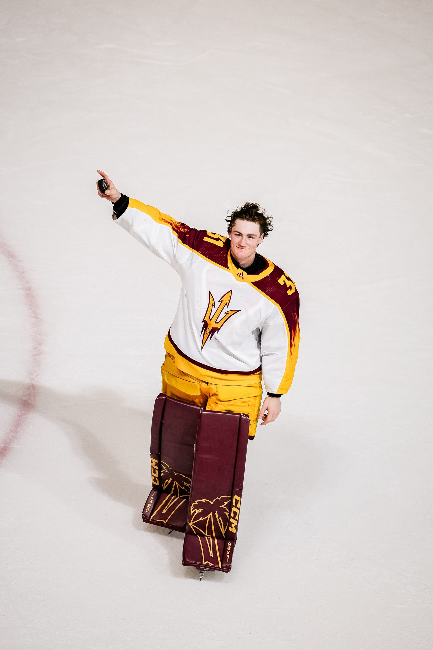 ASU hockey goalie in white and maroon uniform waves to the crowd on the ice rink.