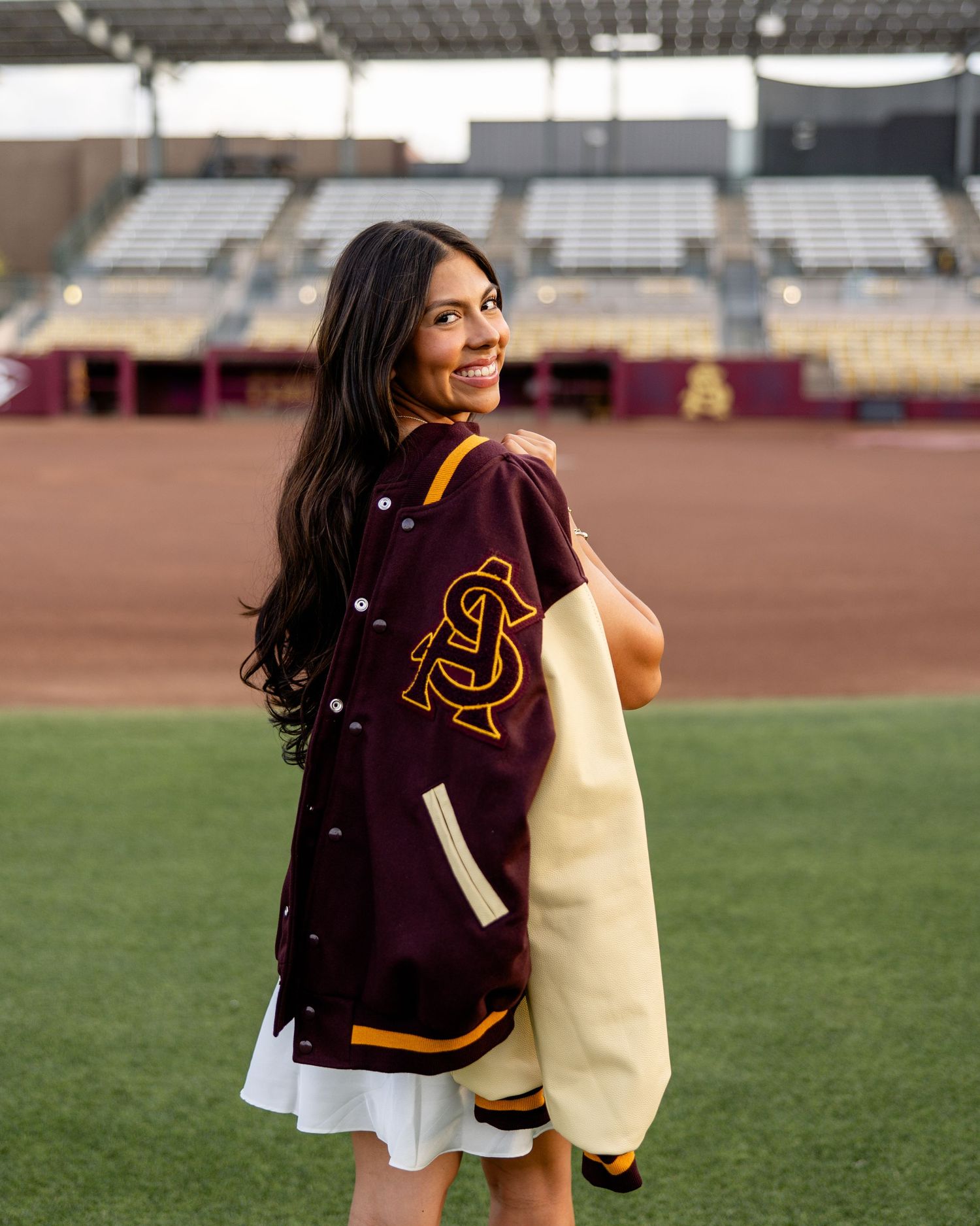 A woman smiles while wearing an ASU maroon and gold varsity letterman jacket on a baseball field.