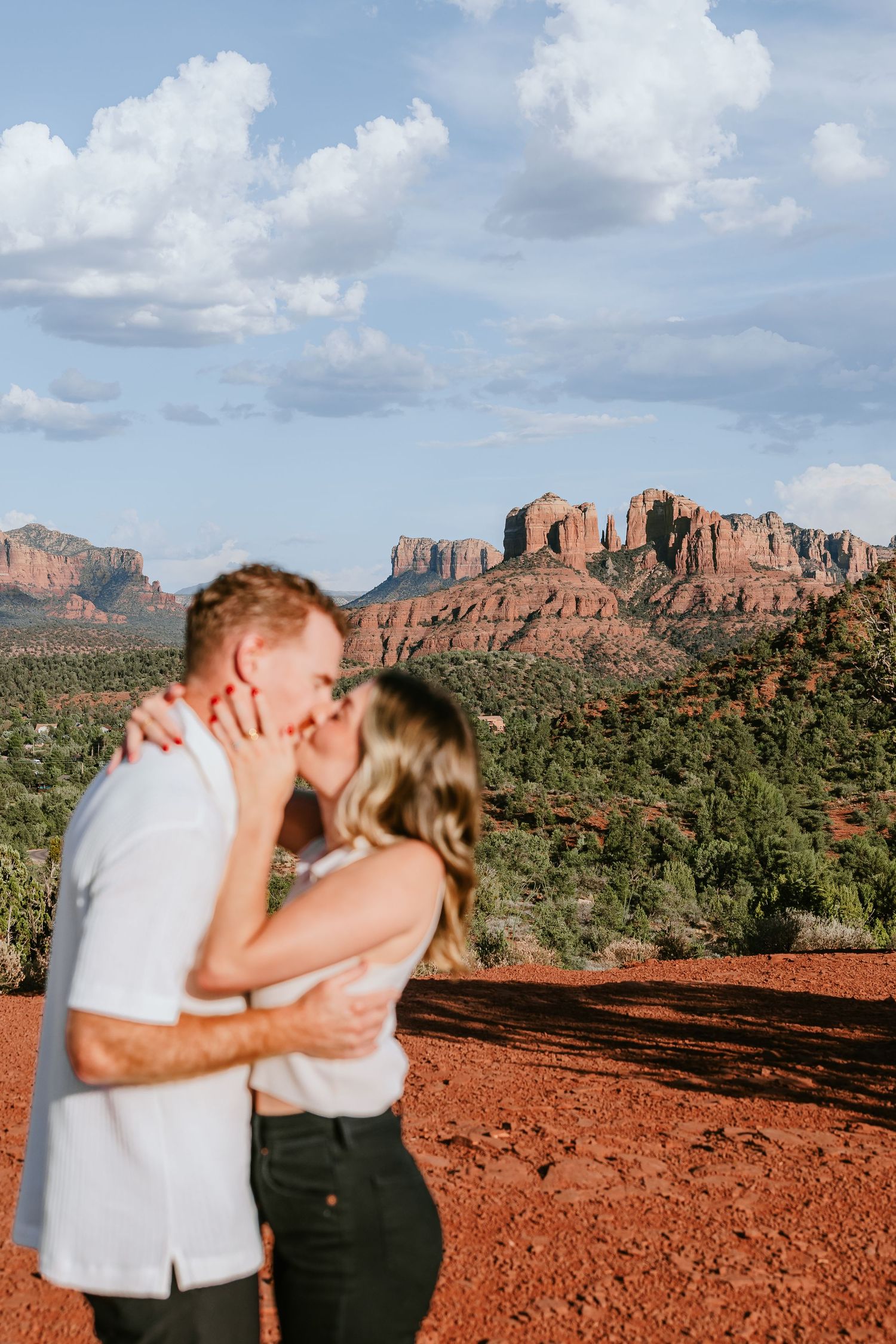 Couple kissing at sunset with Sedona red rock formations and lush green desert landscape in the background.