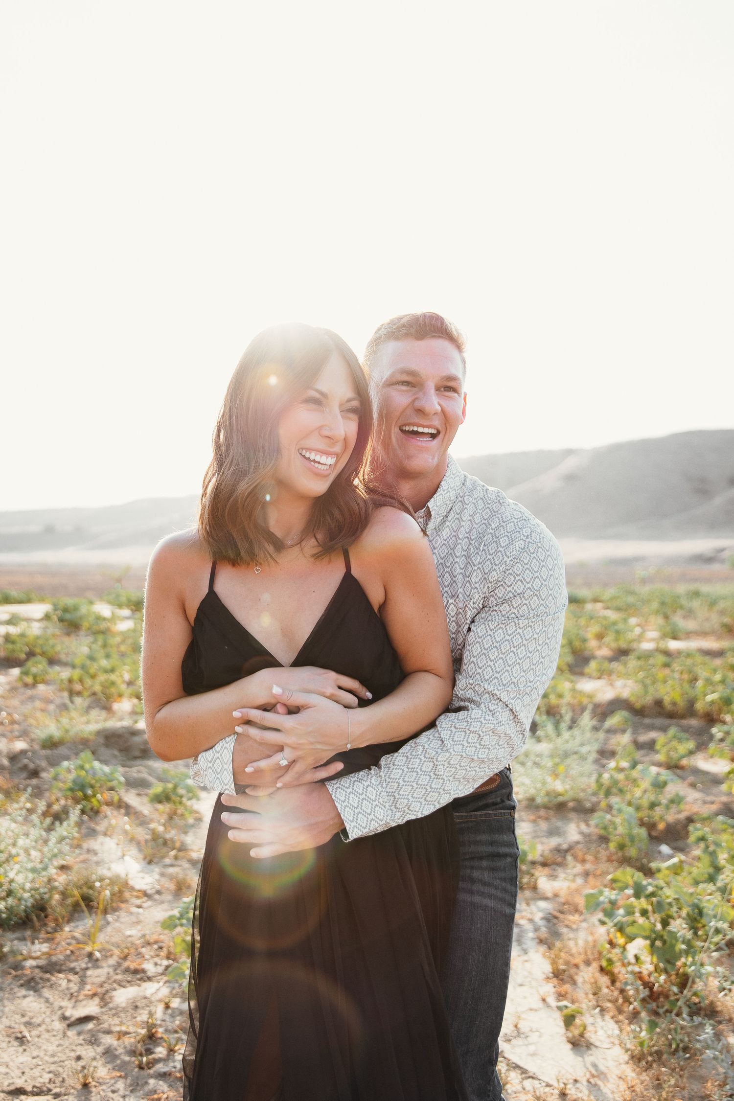 Happy couple embracing outdoors in sunny desert landscape, woman in black dress laughing with man in patterned shirt.