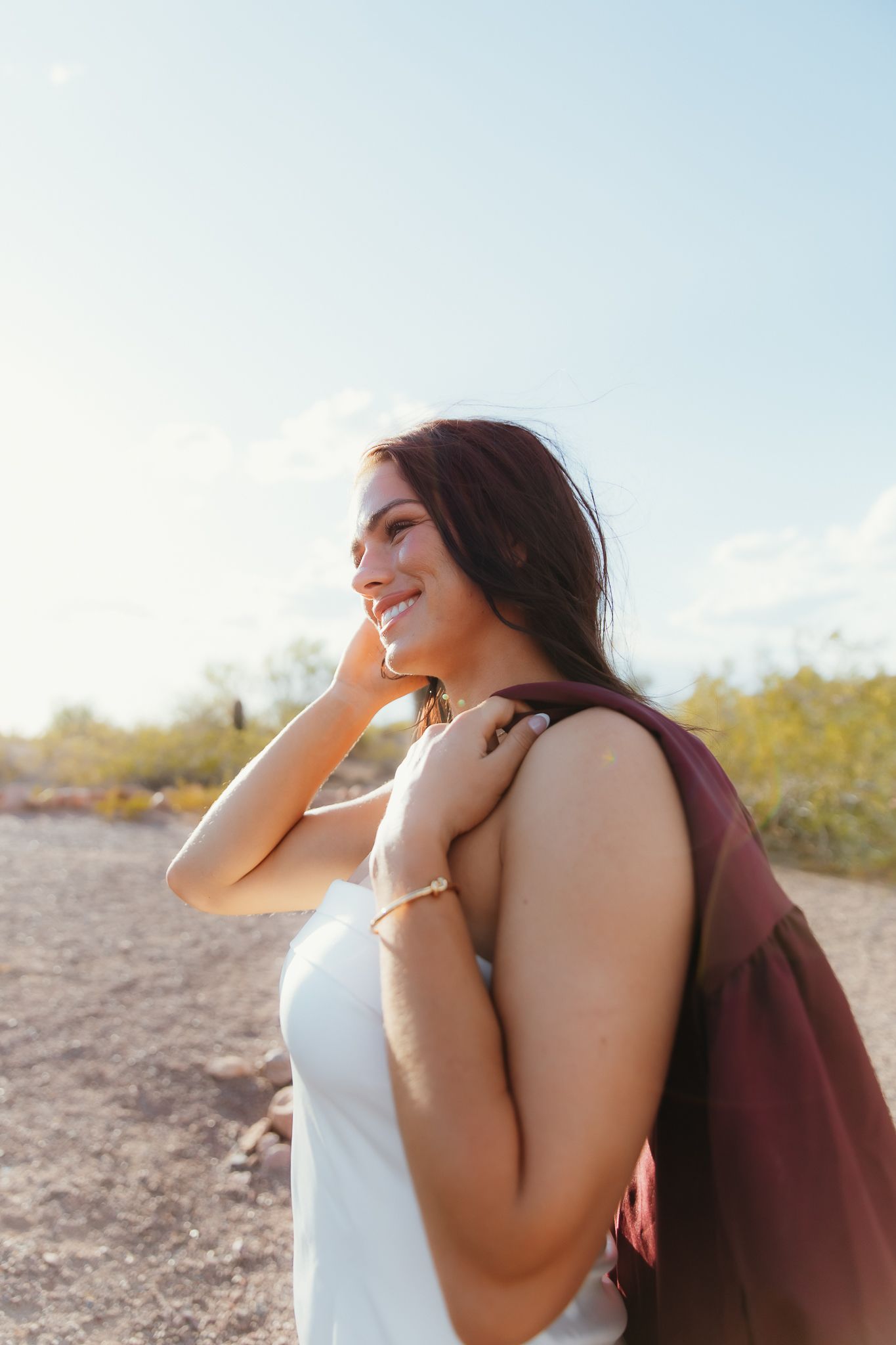 Smiling young woman with dark hair in white dress and burgundy bag poses outdoors in sunny desert landscape.