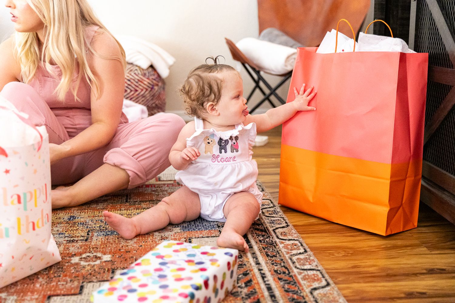 Baby in white romper reaching for a large orange gift bag at a party.