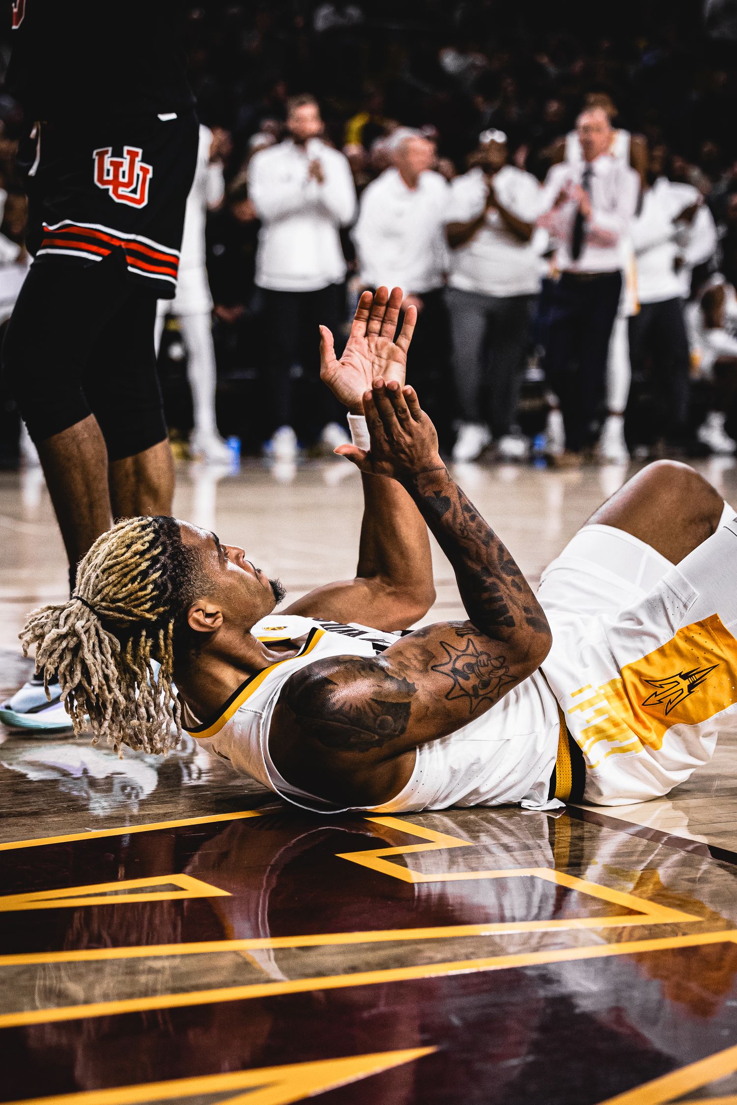 Tattooed Arizona State Sun Devils basketball player lies on the court reaching up during an intense game.