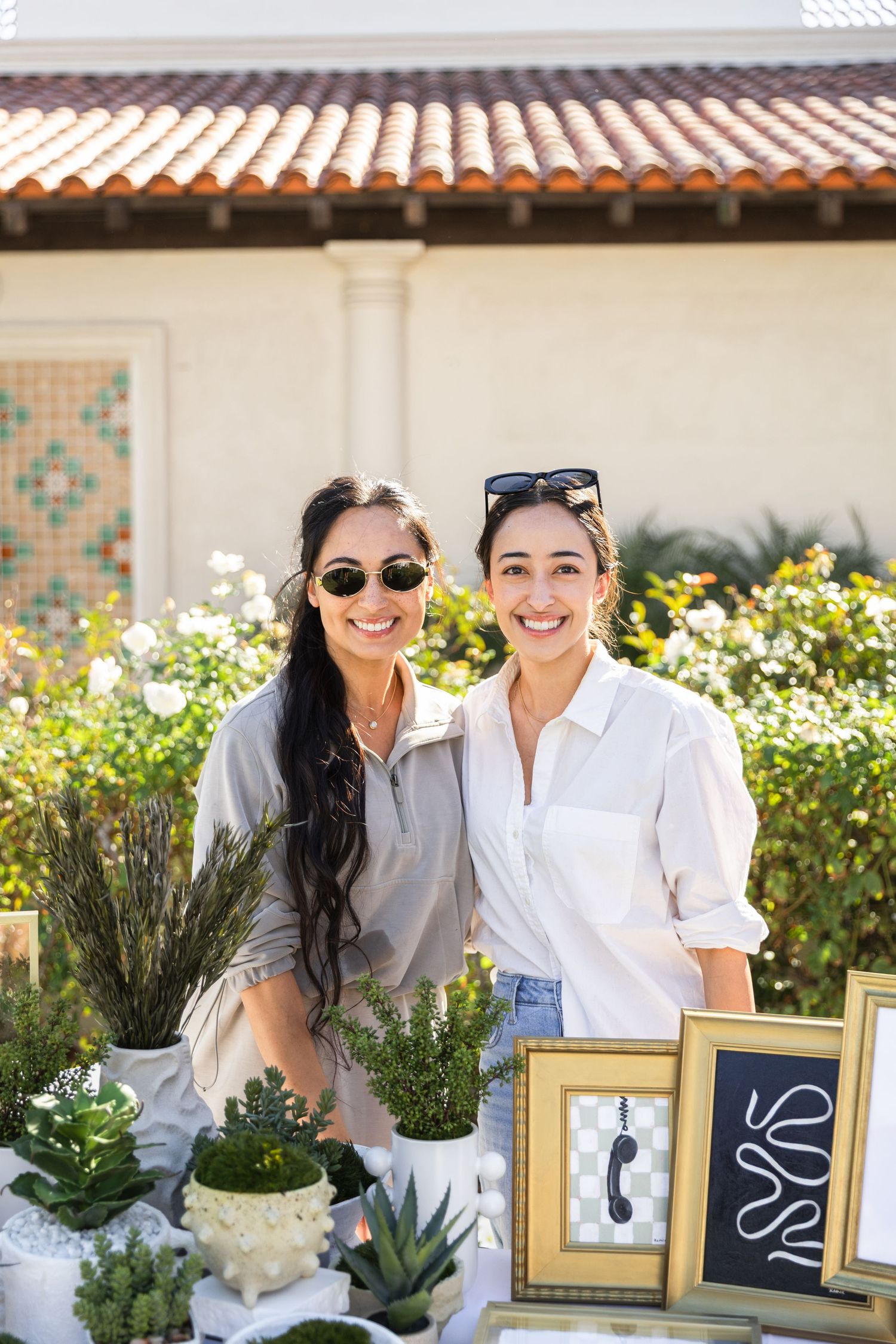 Two smiling women pose behind a table with potted plants and framed art at an outdoor market.