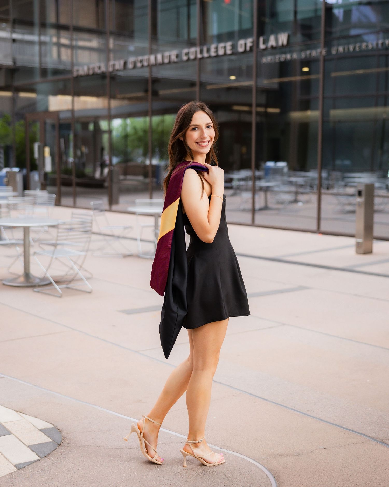 A smiling woman in a black dress carries a maroon graduation hood outside the O'Connor College of Law.