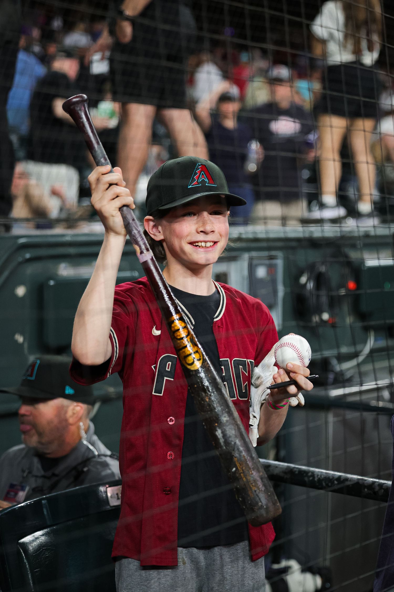 A young fan in an Arizona Diamondbacks jersey smiles while holding a bat and baseball at the ballpark.
