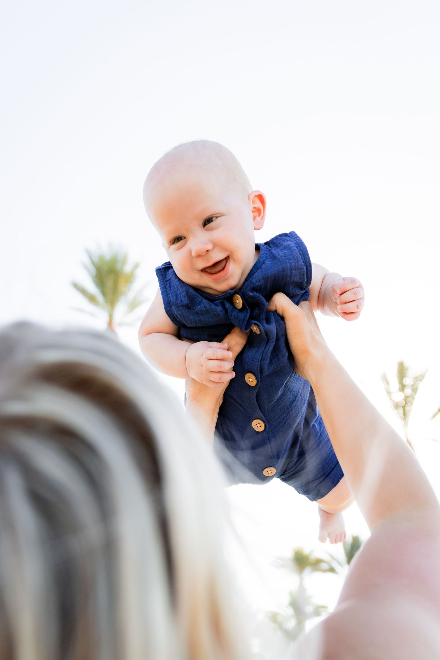 Smiling baby in navy blue romper being lifted up by a blonde mother outdoors.
