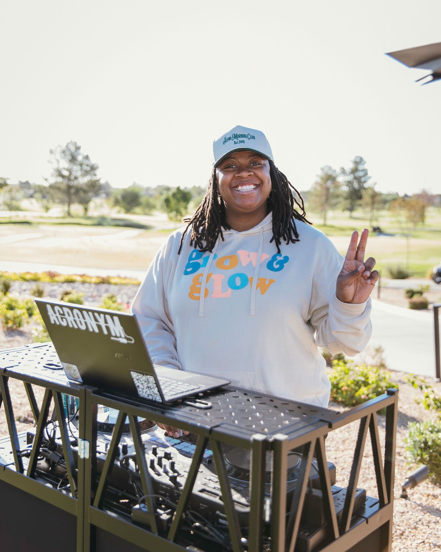 A smiling DJ in a hoodie gives a peace sign while standing behind DJ equipment outdoors.