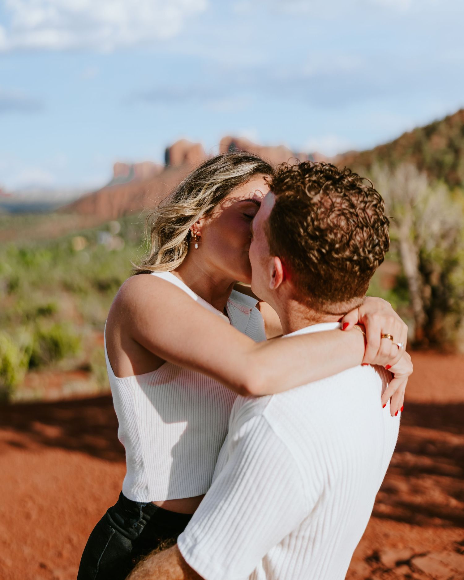 Couple sharing a romantic embrace and kiss in Sedona, Arizona, with red rock formations and desert landscape in background.