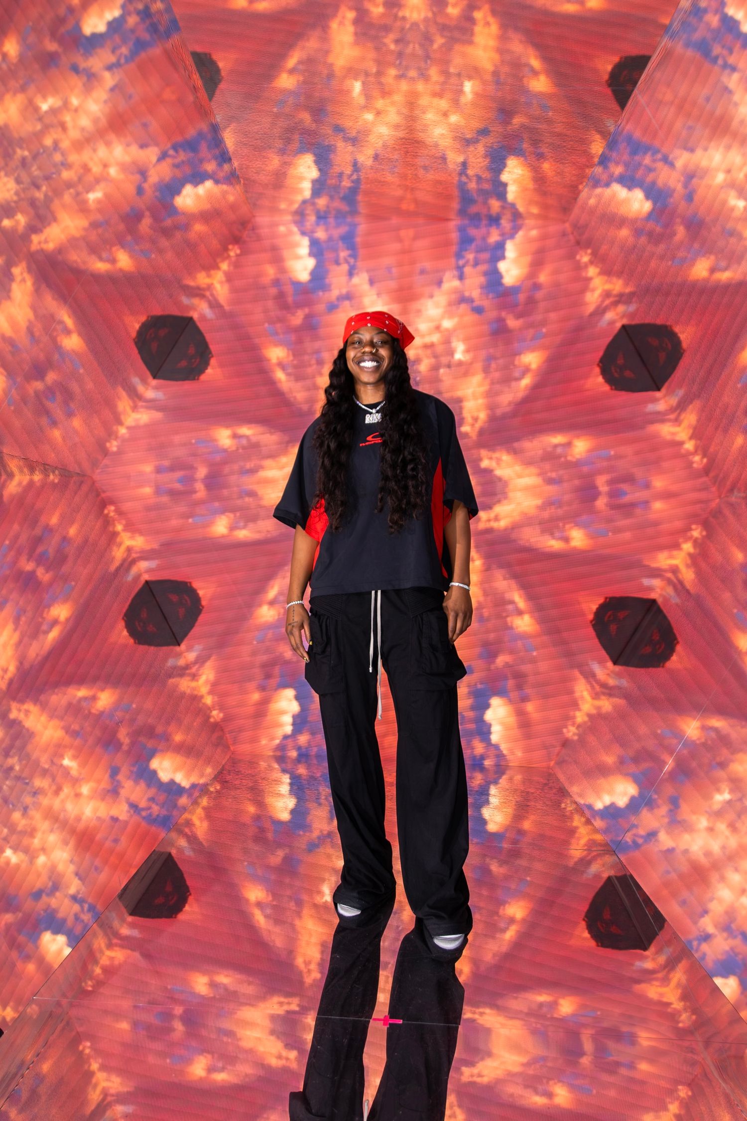 Smiling woman in red bandana and black outfit stands before vibrant red kaleidoscope digital backdrop.
