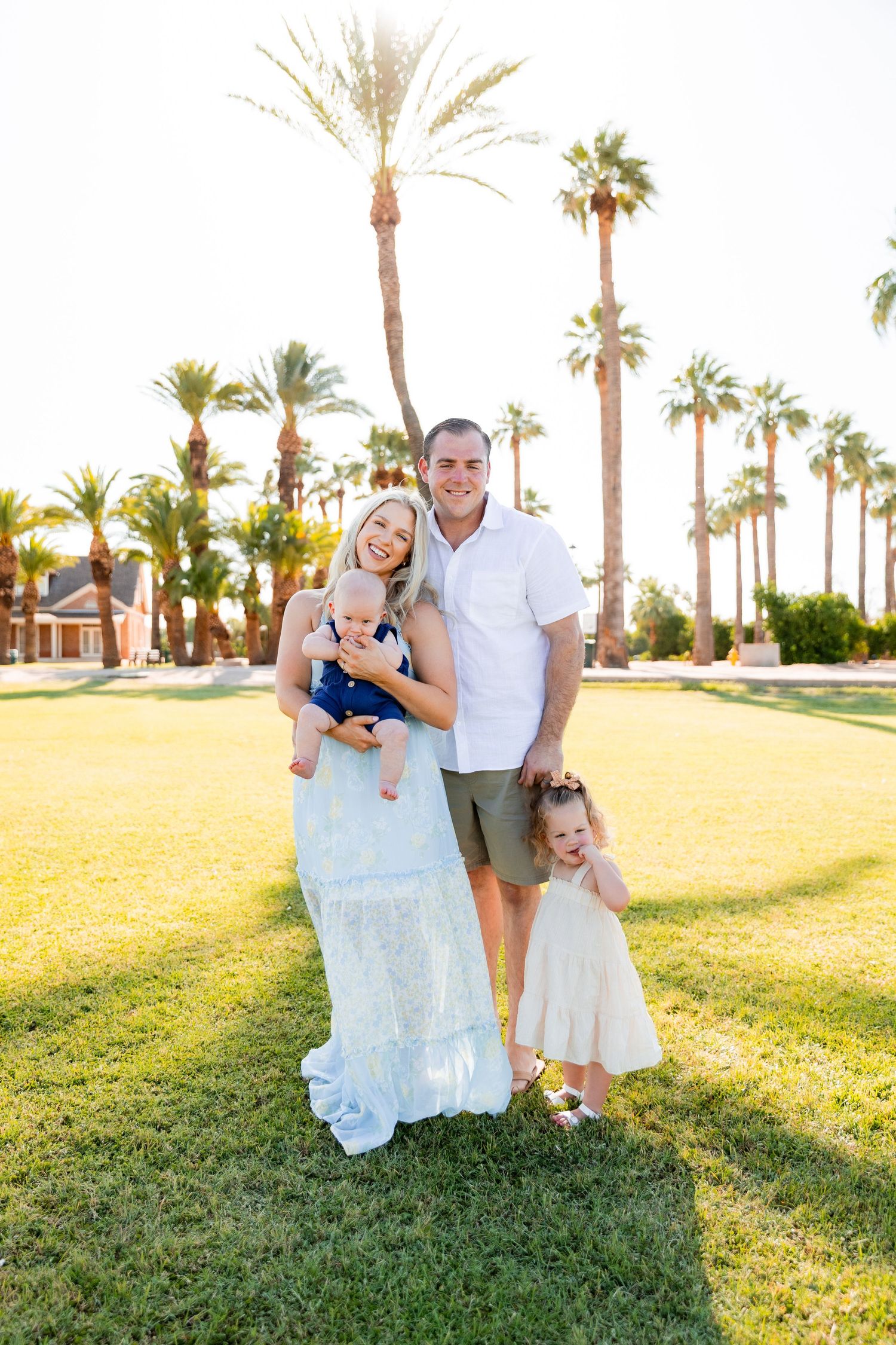 Happy family of four posing on green lawn with tall palm trees in warm golden sunset light.