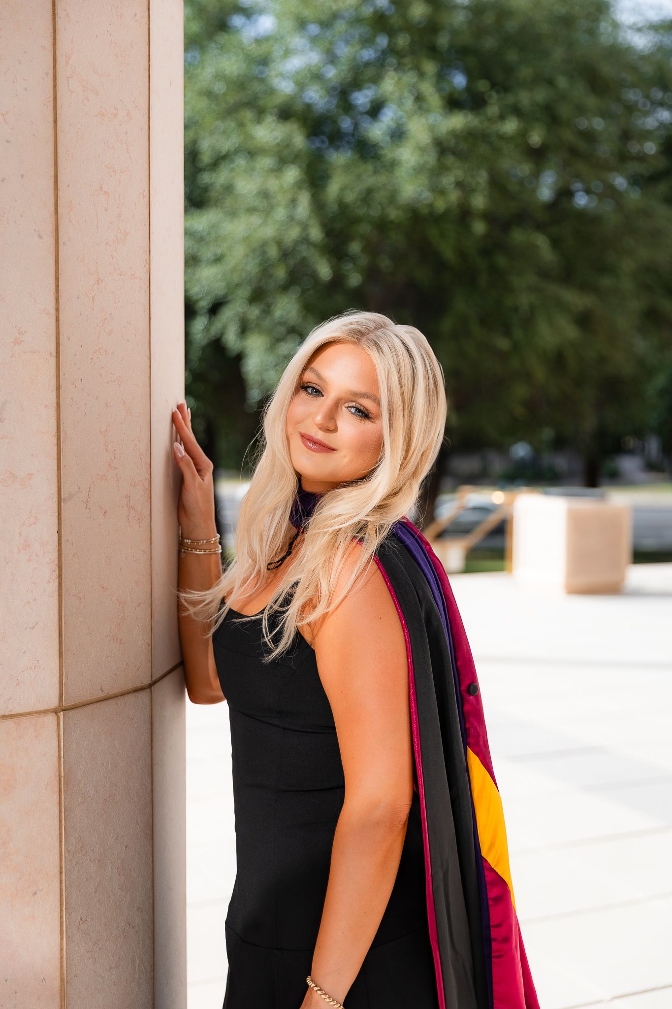 Blonde woman in black dress with graduation stole leaning against a stone column outdoors.