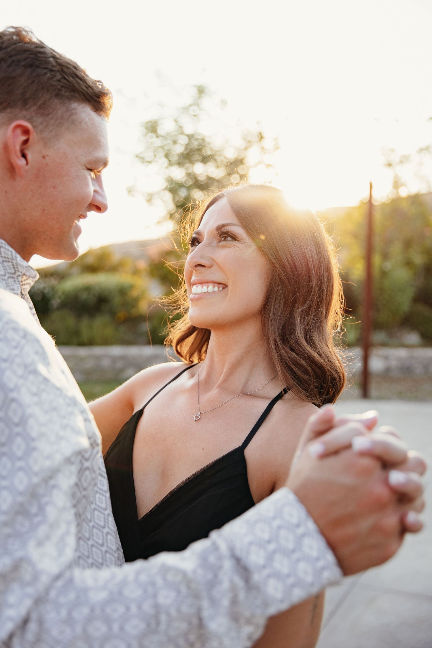 A joyful couple dances outdoors at golden hour, holding hands and smiling at each other in elegant attire.