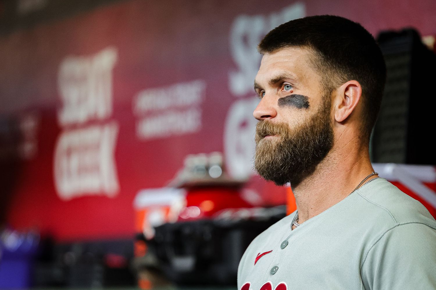 A bearded baseball player with eye black stares intently in a dugout during a game.