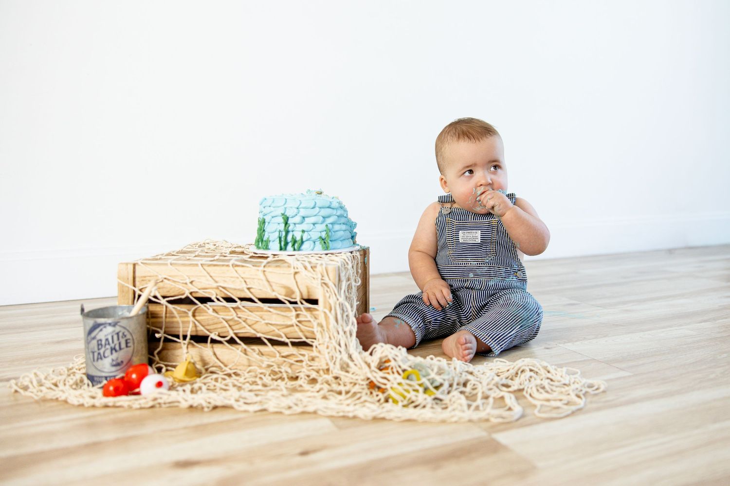 Adorable baby in denim overalls sitting on a rope net beside a wooden crate with colorful toys.