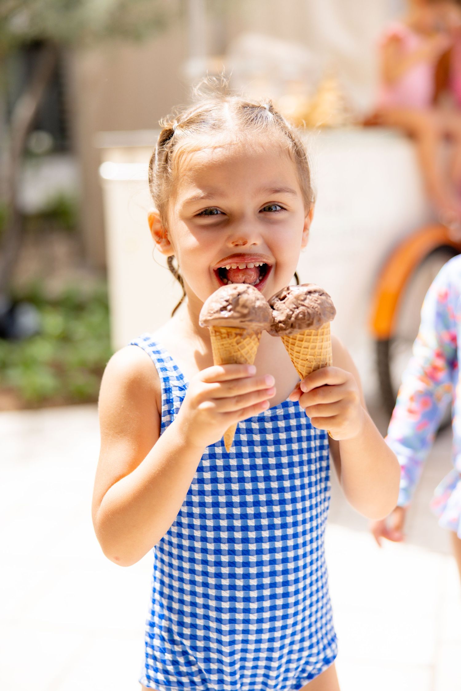 Happy young girl in blue gingham swimsuit eating two chocolate ice cream cones outdoors.